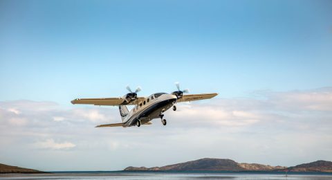 A dynamic photograph of the white and blue Tecnam P2012 Traveller twin-engine commuter aircraft, registration G-OWCY, in motion taking off from the unique beach runway at Barra Airport, External Hebrides, Scotland. This high-wing plane is serving on the vital Stornoway - Benbecula PSO route, showcasing essential connectivity and its STOL (Short Take-Off and Landing) capabilities for the Scottish Highlands and Islands region. Brittle Scottish coastal hills under a partly cloudy sky form the background. This image is for the P2012 entry-into-service press release in Scotland.