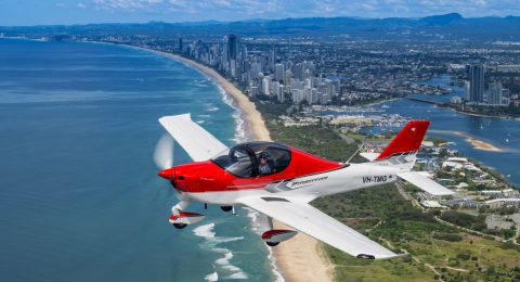 A red and white P-Mentor aeroplane flies above a scenic coastline with tall city buildings, green areas, and a sandy beach alongside the blue sea under a clear sky in Australia.